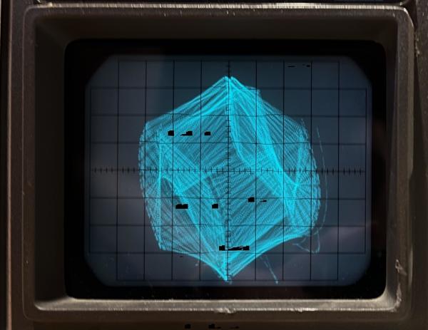 A time lapse of an oscilloscope screen showing an outline in cyan of a cube rotating over time. 