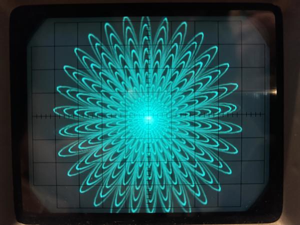 Oscilloscope display in cyan of a repeating flower pattern. 