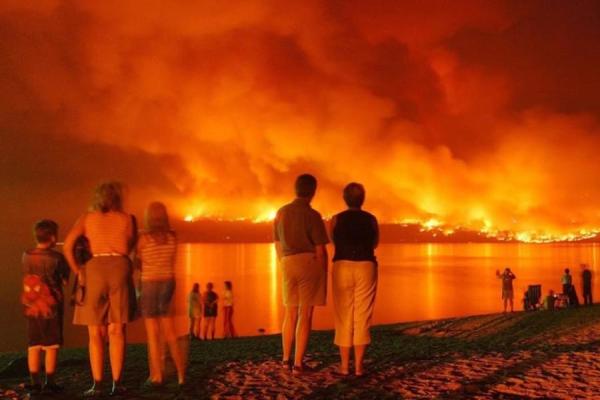 People gathered along a shoreline. They are dressed in summer clothing like shorts and t-shirts. They are watching helplessly as their community burns under a night sky glowing with orange flames and smoke. The image of the fire is reflected in the water of the lake.