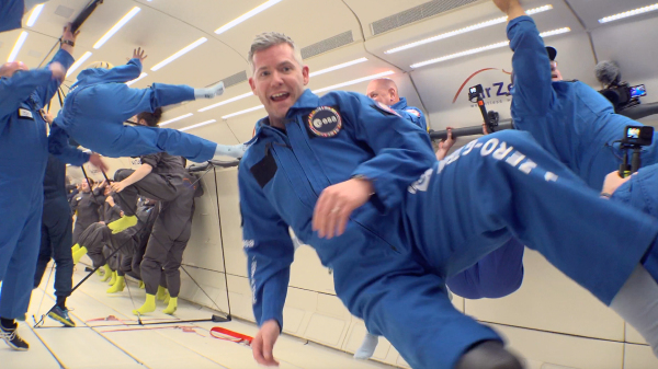 an astronaut in flight suit floats in front of the camera during a parabolic flight. in behind are numerous other astronauts in flight suits spinning in different directions