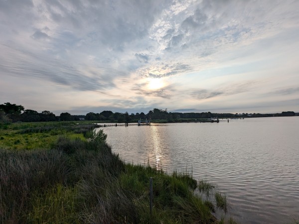 Photo of a calm morning sunrise over Harris Creek yellow sun rising low to the left behind a low broken grey cloud deck. Marsh is green and ready for the day. Creek water is rippled and reflects the sun with a bright sun streak, the sky and clouds.