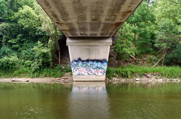 Colourful graffiti tag on the concrete base of a bridge crossing a river as viewed from across the water, the bridge forming a symmetrical roof with River passing underneath and greenery to each side on the far bank. 