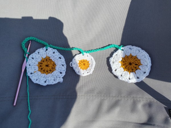 Large crochet daisy, small crochet daisy, large crochet daisy on a chair, all joined together with green yarn. A pink hook is attached on the left