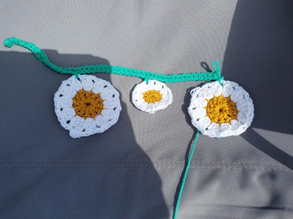 Large crochet daisy, small crochet daisy, large crochet daisy on a chair, all joined together with green yarn with 1 row crocheted