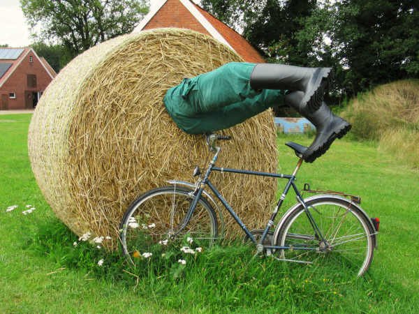 Ein lebensgroßer Dummy steckt mit dem Oberkörper in einem runden Strohballen. Eine gründe Arbeitshose und Gummistiefel ragen fast senkrecht aus dem Stroh. Daran lehnt ein Fahrrad.