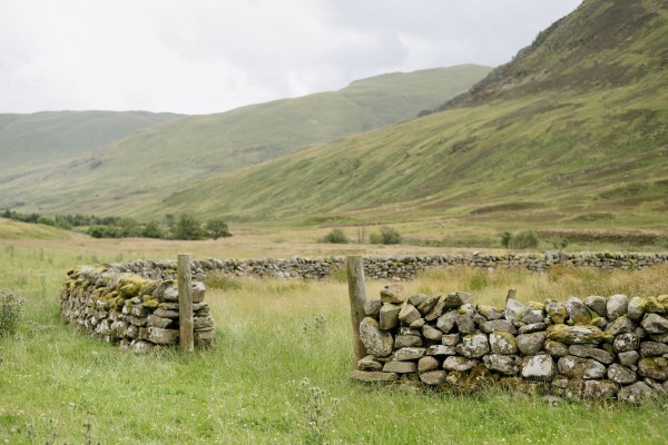 A colour photograph of a circular drystone wall. There is a small gap at the front with wooden posts on either side and all around it hills. 