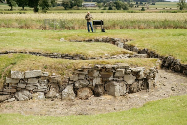 A colpur photograph of a series of drystone walls set into a grassy area. In the distance a man stands beside a sign and behind him a field of barley. 