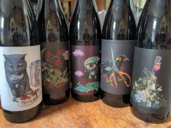 Five large beer bottles with labels depicting 1) a cat with a bowl of bloody food labelled "Mortician" 2) a skull with roses growing out of it 3) a free floating eye surrounded by green goop 4) a black bird carrying a pair of scissors and 5) a scarab beetle clinging to a post surrounded by flowers