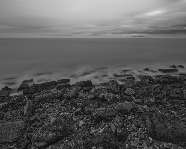 A rocky shoreline, with gentle waves lapping at it. Across the water is a small island with a water tower and industrial-type buildings, somewhat enveloped in fog.