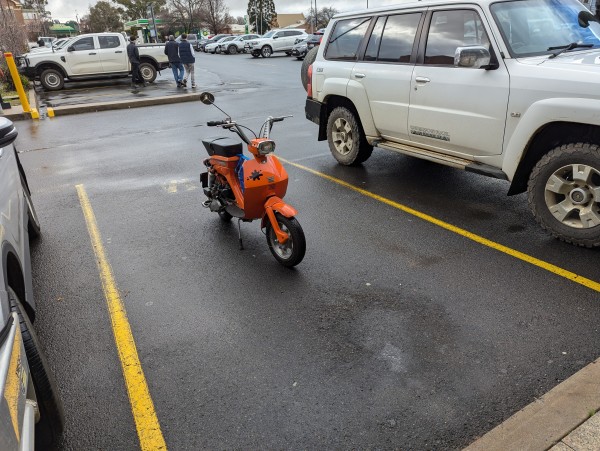 L'il bright orange step through scooter parked in a carpark. It's very angular. 