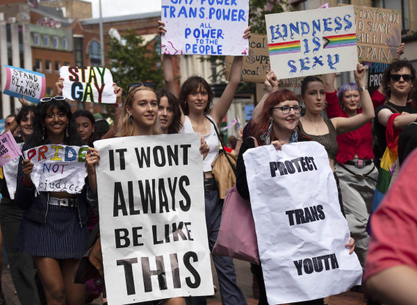 marchers at a rally hold signs aloft and in front of them bearing messages such as "Kindness is sexy", "It won't always be like this" and "Protect trans youth"; buildings in the background