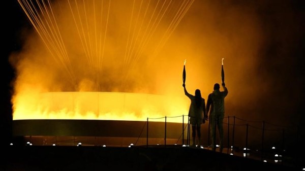 Das Olympische Feuer wurde gerade entzündet. Es befindet sich unter einem riesigen Ballon.
Eine Sportlerin und ein Sportler stehen beide mit einer Fackel davor.