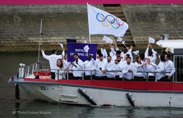 Group of people on a boat holding a flag with the Olympic rings and a banner that reads "Refugee Olympic Team."