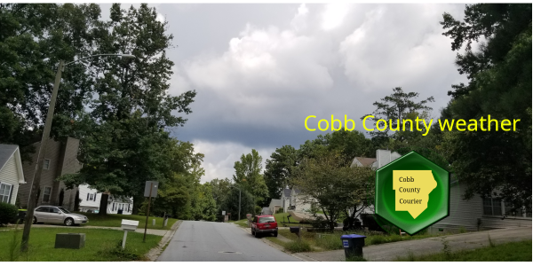 A Cobb County residential street under cloudy skies with detached houses and parked cars to the side, overlaid with a Cobb County Courier logo and the words "Cobb County Weather"
