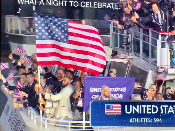 A photograph showing a large group of people on a boat, enthusiastically waving American flags. Lebron James in a light-colored jacket is prominently holding a large American flag in the foreground. The crowd appears to be celebrating, with smiles and raised flags. In the background, a banner reads "UNITED STATES OF AMERICA." The overlay text at the top of the image reads "WHAT A NIGHT TO CELEBRATE." At the bottom right corner, there's an overlay box with the text "UNITED STATES" alongside the American flag icon and "ATHLETES: 594."