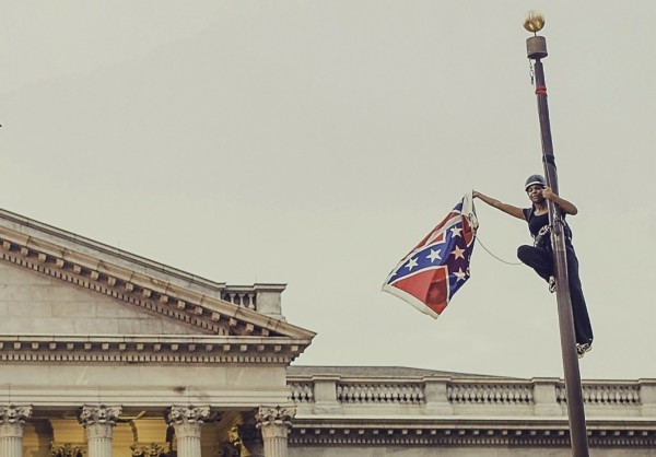 Bree Newsom at the top of the flagpole, with the removed flag in her hand. She is a black woman in a climbing rig and hard hat.