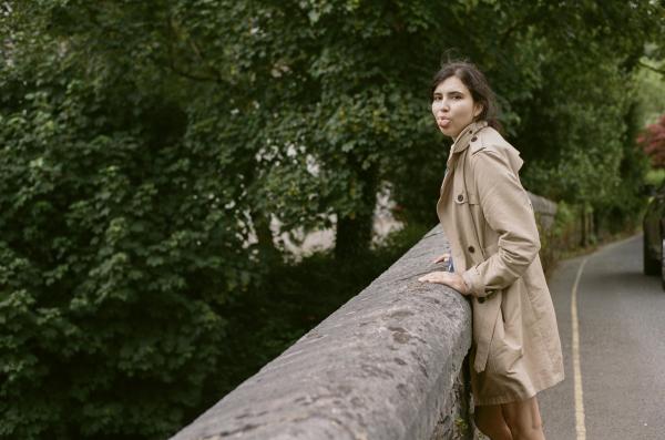 A woman with dark hair, wearing a beige trench coat, leans on the stone edge of a bridge, sticking her tongue out mockingly to the camera man. 