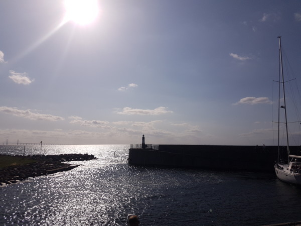 Ein Boot liegt vor einer Mauer, hinter der die offene See beginnt. Die Sonne geht unter. 