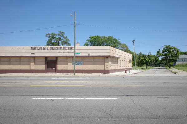 Color photograph looking across a wide empty street at a broad tan church building, windows and doors along the street level are all boarded over, and there's somewhat fading handpainted text above the entrance, "New Life M. B. Church of Detroit." The building is right up against the sidewalk and wraps around a corner with an obtuse angle