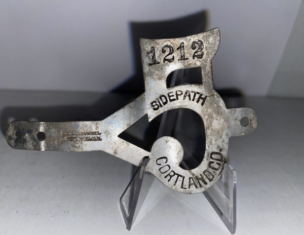 a metal band, meant to be wrapped around a bicycle fork frame, with "Sidepath" and the number 1212 and "Cortland Co" stamped into it.  The entire band is cut intot the shape of the numeral 5.