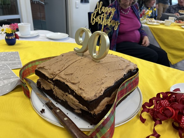 A large square cake sitting on a plate on a table. It’s dark chocolate, with a lighter brown creamy chocolate filling and icing. On top are large candles with the numbers 90 and a sign saying Happy Birthday. A red tartan ribbon and red frill that were around the cake have been removed and the first cut made.