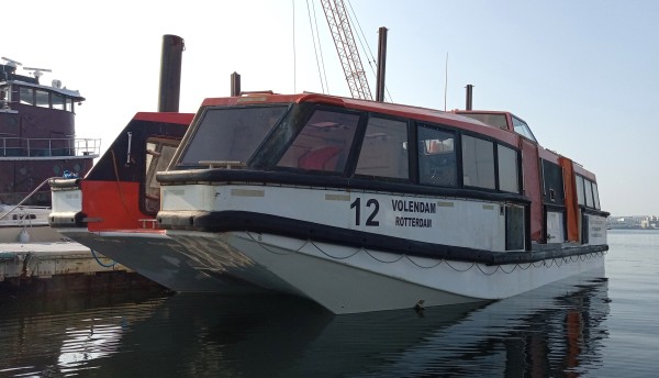 Photo of a blocky-looking large entirely enclosed lifeboat, tied up at a dock near an old tugboat.