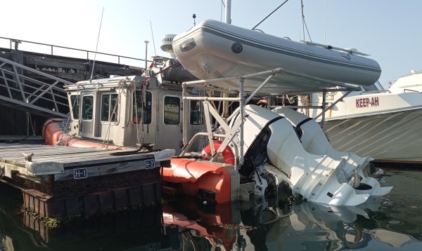 Photo of the heavy-duty lighthouse tender Gerda, showing her enclosed cabin, dual outboards, tender hanging from a mast, and a substantial chunk ripped out from the stern of her orange hull.