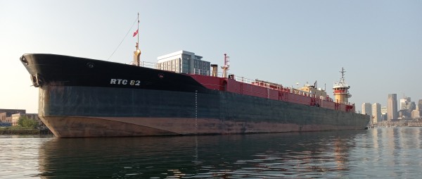 Photo of the bow of a large oil barge with a black hull and red topsides. You can see the pusher tug on the barge's stern and a glimpse of the downtown Boston waterfront across the harbor.