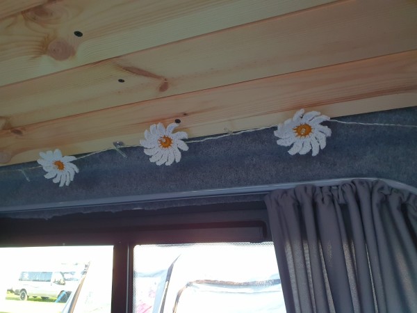 3 crochet daisies poked onto a string of fairy lights running along a tongue and groove ceiling. Below the ceiling is grey carpet and part of a window which has a grey curtain on it