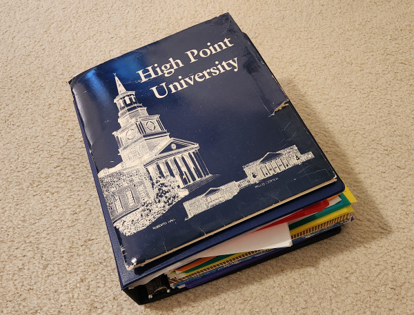 A collection of binders and folders containing college coursework. The top binder, supplied by the college, has the name "High Point University".