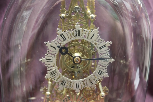 A skeletonized clock where you can see all the gears and wheels. The clock face is a silvered ring with black Roman numbers. The clock movement is sitting under a dome