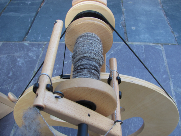 A modern spinning wheel in light-coloured wood seen from above. The bobbin is stationary, partly full of fine grey yarn which echoes the mottled grey of the slate flags on which the spinning wheel stands