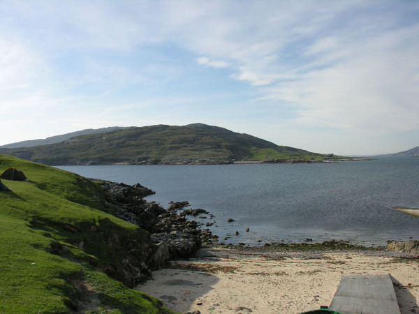 Scarp in the Western Isles. The image shows a beach and concrete slipway in the bottom right of the frame with rising grassy ground on the bottom left. There’s a small tent in the left centre of the frame. Then there is a stretch of sea with, beyond it, a low lumpy island. The scene is in sunlight.