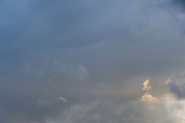 Photo of a bit of a large blue-grey cloud mass, varying little across the picture, other than: in places across the lower third smaller clouds catch slivers of light; at the bottom-right corner a small puff catches s