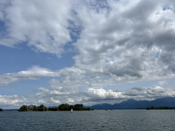 Inmitten des Chiemsees. In der Ferne die Krautinsel, zwischen Frauen- und Herreninsel liegt. Davor ein kleines Segelboot. Am rechten Bildrand die Chiemgauer Berge.  