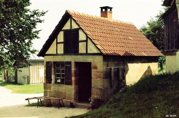 Farbfoto eines Backhauses im Freilichtmuseum Beuren auf der Schwäbischen Alb. Es handelt sich um ein größeres Gebäude in Stein- und Fachwerkkonstruktion mit Eingangstür, Fenster und steilem Ziegeldach. Vor dem Haus befinden sich zwei primitive Holzbänke. Rechts ein grasbewachsener Hügel, links ein Weg, ein weiteres Gebäude und ein Baum. Leica R5 mit 50 mm Summilux auf Adox Color Implosion.