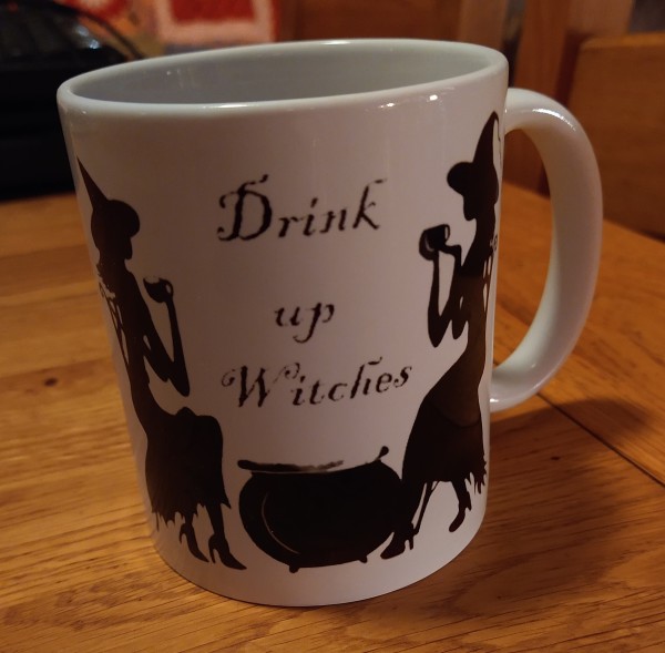 Mug of tea on a dining table, it has two witches sitting either side of a cauldron. Sipping tea, the words 'Drink up Witches' in the centre 