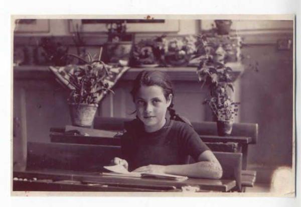 Vintage black and white photo of a girl seated at a wooden desk inside a classroom, writing in a notebook. There are several potted plants on a shelf behind them.