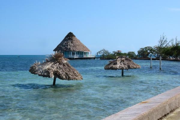 Photo of a submerged beach, with two palm thatched umbrellas sticking out of the sea. In the foreground there is part of a concrete sea wall visible, and in the background, a thatched cabana. The sea is a beautiful turquoise and blue, under a clear blue sky.