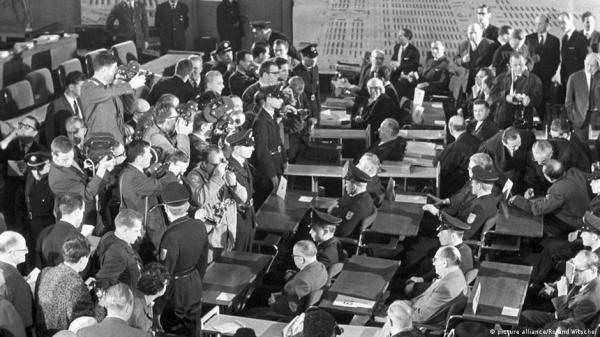 Black and white photo of a group of journalists with cameras and notepads, attentively covering an event of a trial in a formal setting filled with seated attendees.