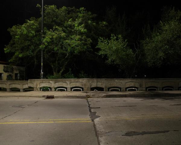 Color photograph looking across a street on a bridge at night, the sidewalk opposite broken up in some places alongside a thick, short concrete barrier. Green leaves and branches of trees grown up from below the bridge stretch out under the light of a streetlight, illuminated against the darkness behind them