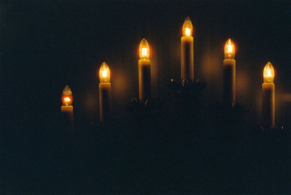 A grainy analog photograph of electric Advent lights in a windowsill (you'll just have to trust me on the windowsill part). The picture is exposed for the small and glowy, incandescent lightbulbs, so everything around is just black. The film stock creates some slight halation around the light bulbs, and there is a blurry reflection in the matte window behind the lights.