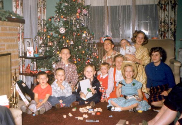 Eleven children open presents in front of a live Christmas tree, posed for a group photo. One boy received wooden block. One girl wears a cowboy hat, gun belt and twirls a sixshooter. No one else shows their presents.