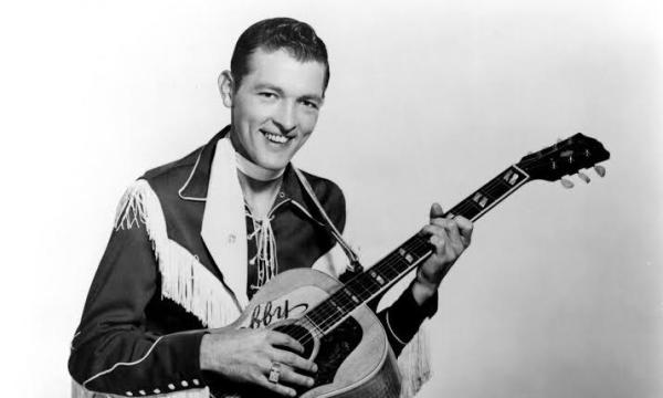 Bobby Helms, en una fotografía en blanco y negro tocando su guitarra con cara sonriente y un traje de vaquero negro con flecos blancos. Dominio público.
