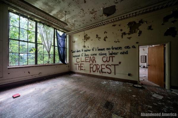 An empty classroom with a wooden floor, peeling paint, and worn drapes on the windows. On the wall someone has spray painted, "what does it matter if i leave no trace as a boyscout if they come along later and clear cut the forest"