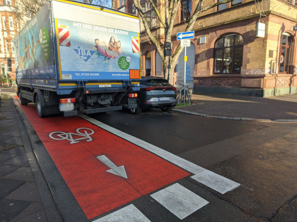 Jemand fährt mit einem LKW in einer recht schmalen Straße in Gegenrichtung über einen Radfahrstreifen, rechts davon ein geparktes Auto am Fahrbahnrand