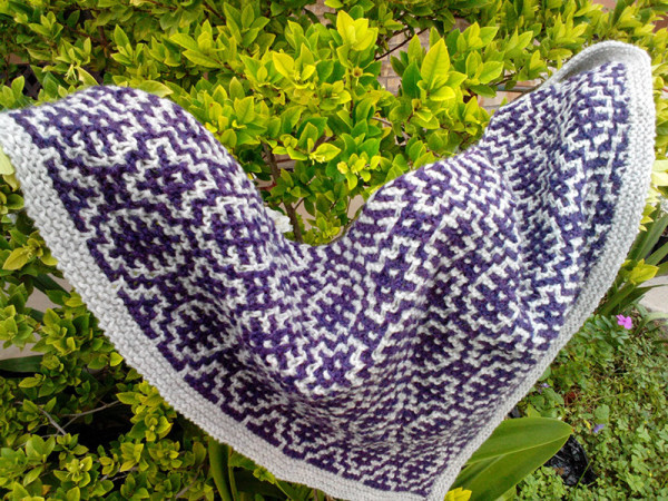 A knitted shawl consisting of mosaic garter colorwork. The main body is in pale grey while the motifs are in a plum purple. 