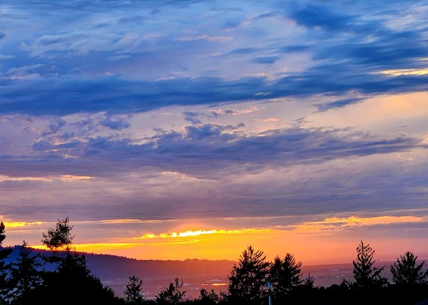 Cloudy sky with dark blue clouds. Gold where the sun is closer to the horizon. Fog just forming in the valley. Dark tree tops at the bottom.