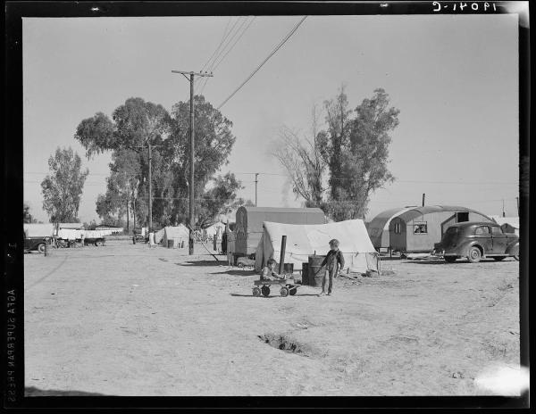 The image is a black and white photograph depicting an outdoor scene with several makeshift tents or shacks lined up in rows. There are telephone poles visible on the left side, suggesting that this might be near a road or power lines. Amongst these temporary dwellings, there appears to be a small child seated on what looks like a toy wagon pulled by another individual.

In front of one of the shelters, two young boys stand beside each other; they seem engaged in some activity with objects on the ground. The setting is an open dirt field under a clear sky, and beyond the tents are trees that appear to be eucalyptus or similar species, which indicates this might be near California.

A vintage car parked at one end of the line suggests that it's likely from mid-20th century America. There seems to be some text on top of the image but it is not clearly legible in this context due to resolution constraints and angle perspective issues within the photo frame; however, there appears a possible watermark or note reading '1948.'