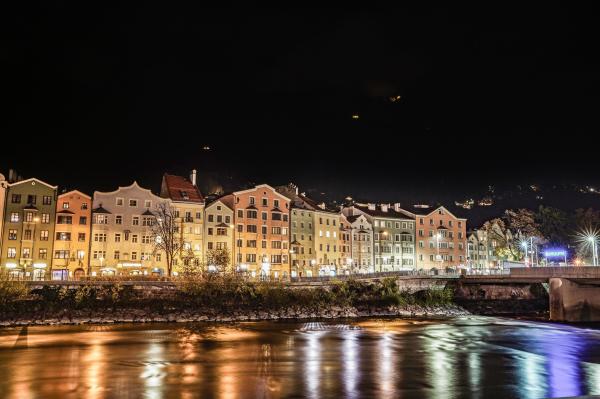 Das Foto zeigt eine nächtliche Szene der Mariahilf-Reihe in Innsbruck, Österreich. Die charakteristischen farbigen Häuser entlang des Flusses Inn bilden das zentrale Motiv. Die Fassaden der Gebäude, in zarten Pastellfarben gehalten, sind von warmem Licht angestrahlt, wodurch sie sich eindrucksvoll vom dunklen Nachthimmel abheben.

Im Vordergrund fließt der Inn ruhig und reflektiert die Lichter der Häuser und Straßenbeleuchtung, was ein faszinierendes Farbenspiel auf der Wasseroberfläche erzeugt. Der Fluss wird von einer Brücke am rechten Bildrand überspannt, und nahe der Brücke leuchtet ein Schild mit der Aufschrift „INNRAIN“, das auf die dortige Straße hinweist.

Im Hintergrund sind die dunklen Silhouetten der Berge zu erkennen, mit einigen schwach leuchtenden Lichtpunkten, die auf Hütten oder andere Gebäude in der Höhe hinweisen. Die Szene kombiniert städtische Eleganz mit der natürlichen Schönheit der umgebenden Berglandschaft und vermittelt einen Eindruck von Ruhe und Harmonie in der Nacht.